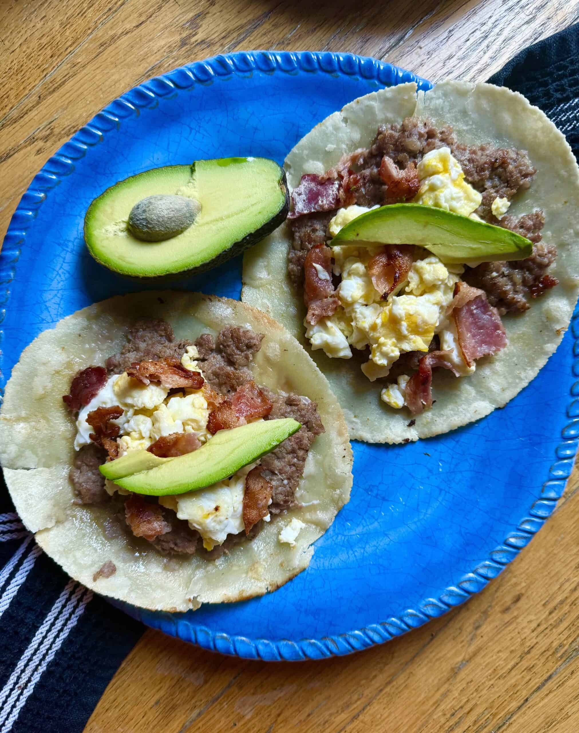 Two breakfast smashed tacos on almond flour tortillas with ground sausage, scrambled eggs, bacon, and avocado slices