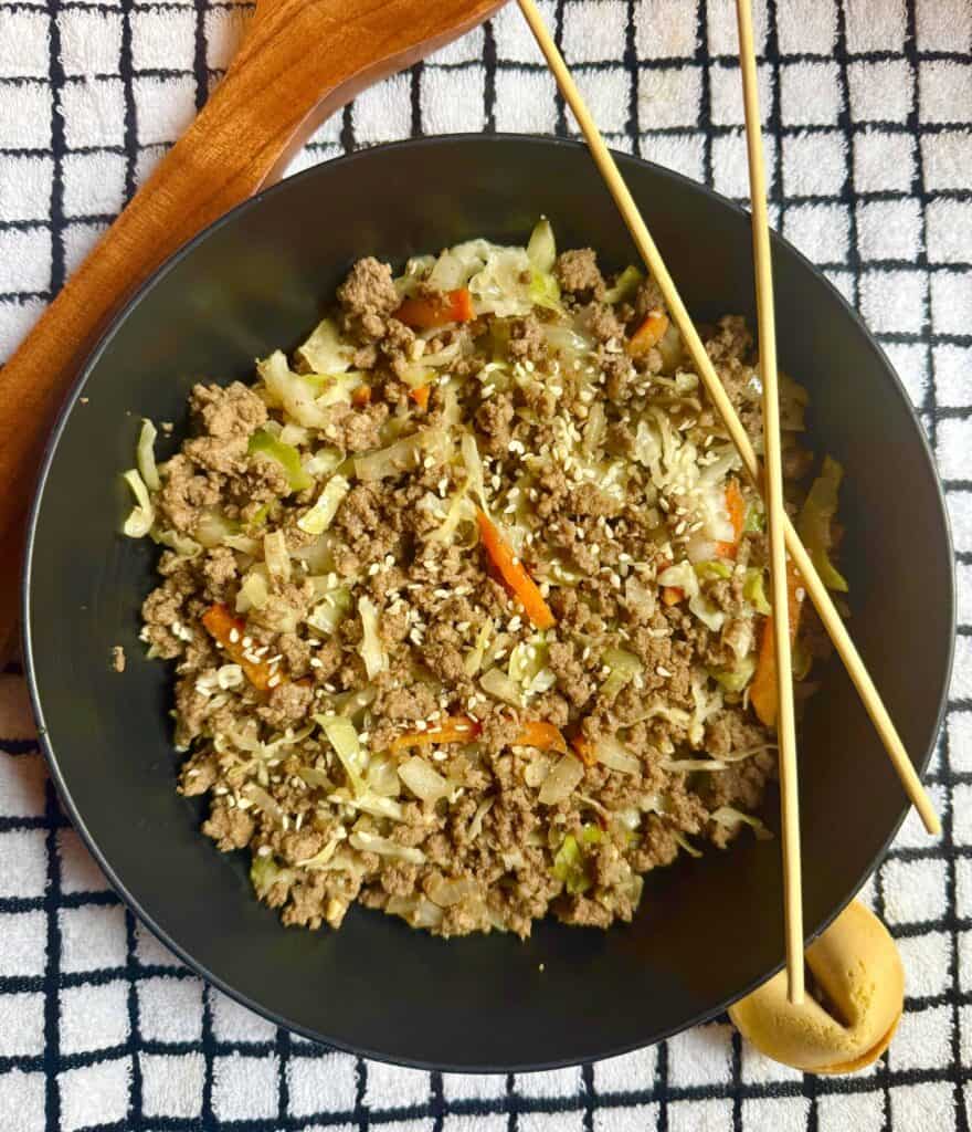 Ground beef egg roll in a bowl served in a black bowl, made with sautéed cabbage, onions, carrots, and sesame seeds, photographed overhead on a white grid towel with chopsticks.