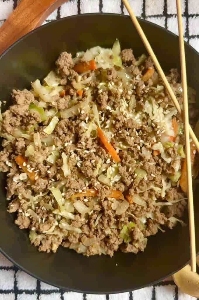 Overhead view of a low-carb egg roll in a bowl with ground beef, cabbage, onions, carrots, and sesame seeds, styled in a black bowl with wooden chopsticks on a checkered kitchen towel.
