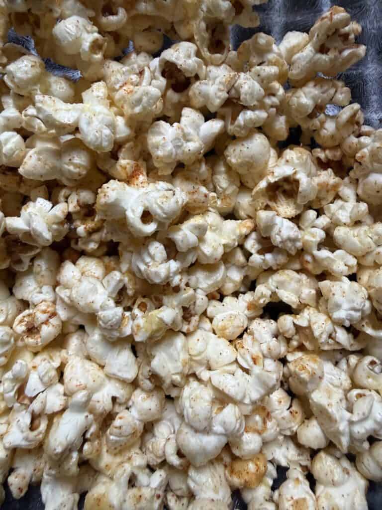 Close-up of elote popcorn coated with melted butter, Parmesan cheese, and a light chili seasoning, showing individual popped kernels with visible spice.

