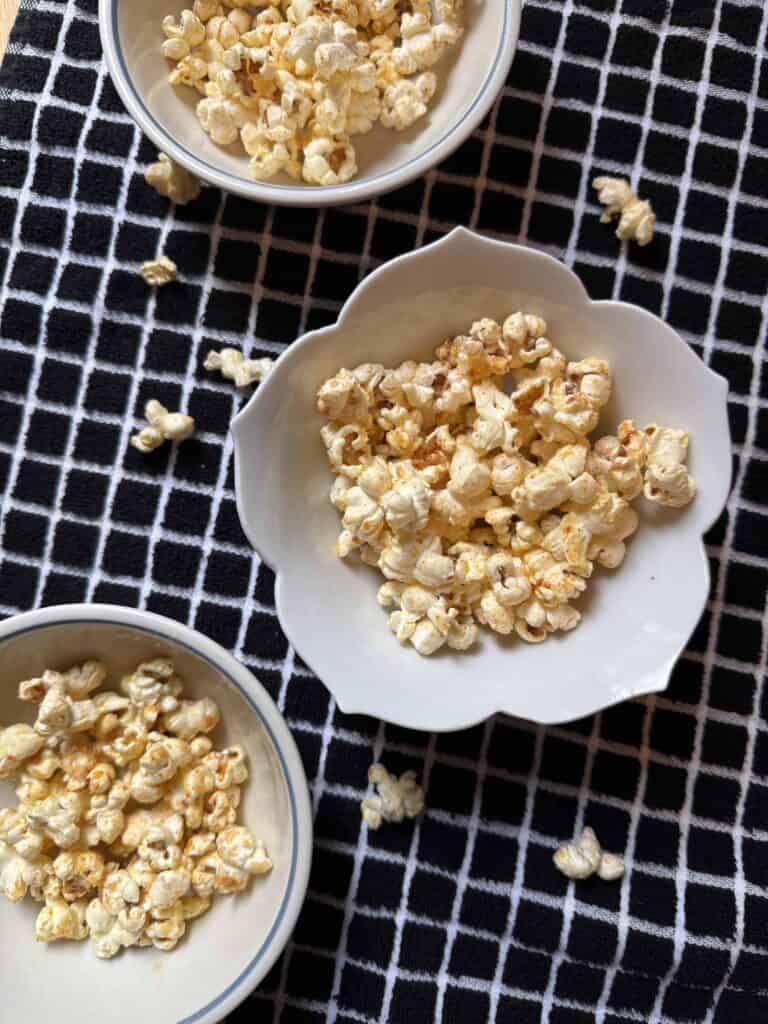 Overhead view of elote popcorn served in small white bowls on a black-and-white kitchen towel, seasoned with butter, Parmesan cheese, and chili spices.