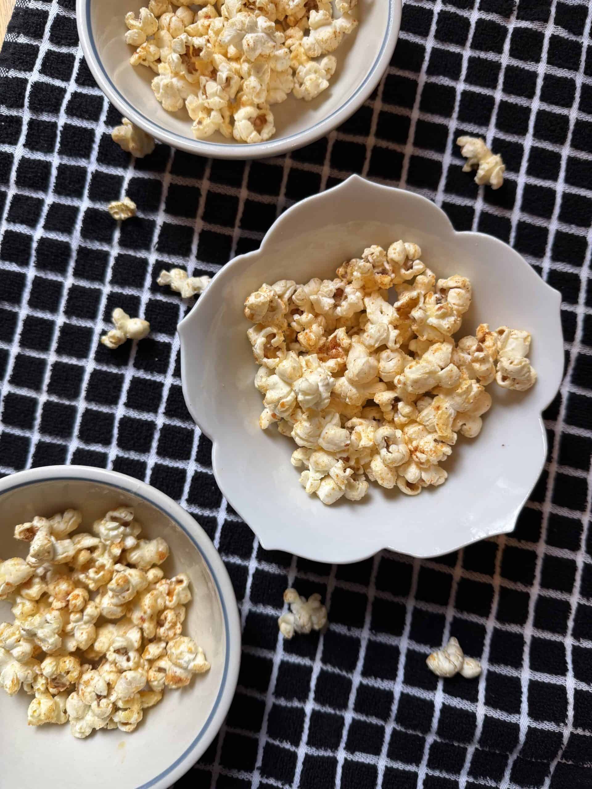 Overhead view of elote popcorn served in small white bowls on a black-and-white kitchen towel, seasoned with butter, Parmesan cheese, and chili spices.