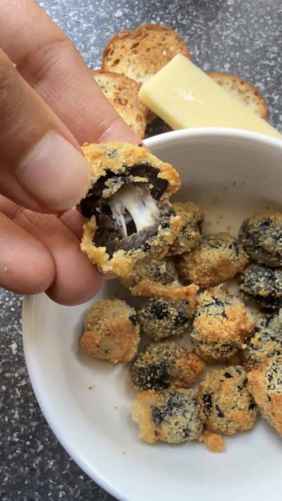 Close-up of a crispy breaded black olive with melted cheese stretching from the center, held between fingers, with more baked olives in a bowl in the background.
