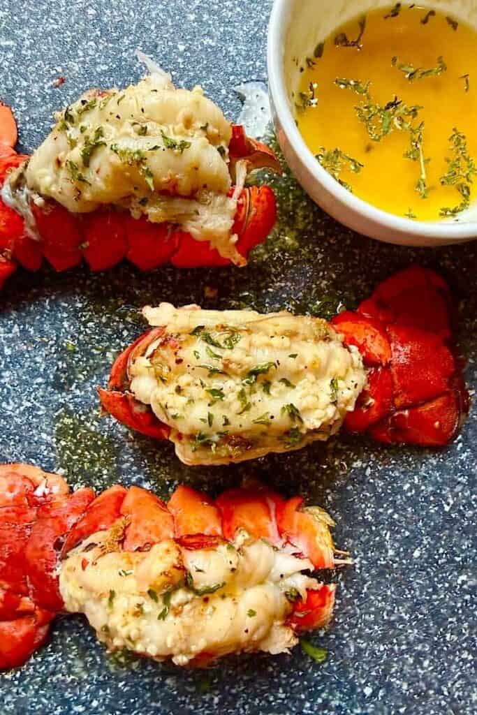 Broiled garlic butter lobster tails served on a blue plate with a small bowl of melted garlic herb butter on the side.