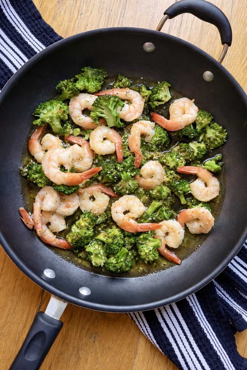 Black nonstick skillet filled with sautéed shrimp and broccoli in a light garlic sesame sauce on a wooden table with a striped kitchen towel.