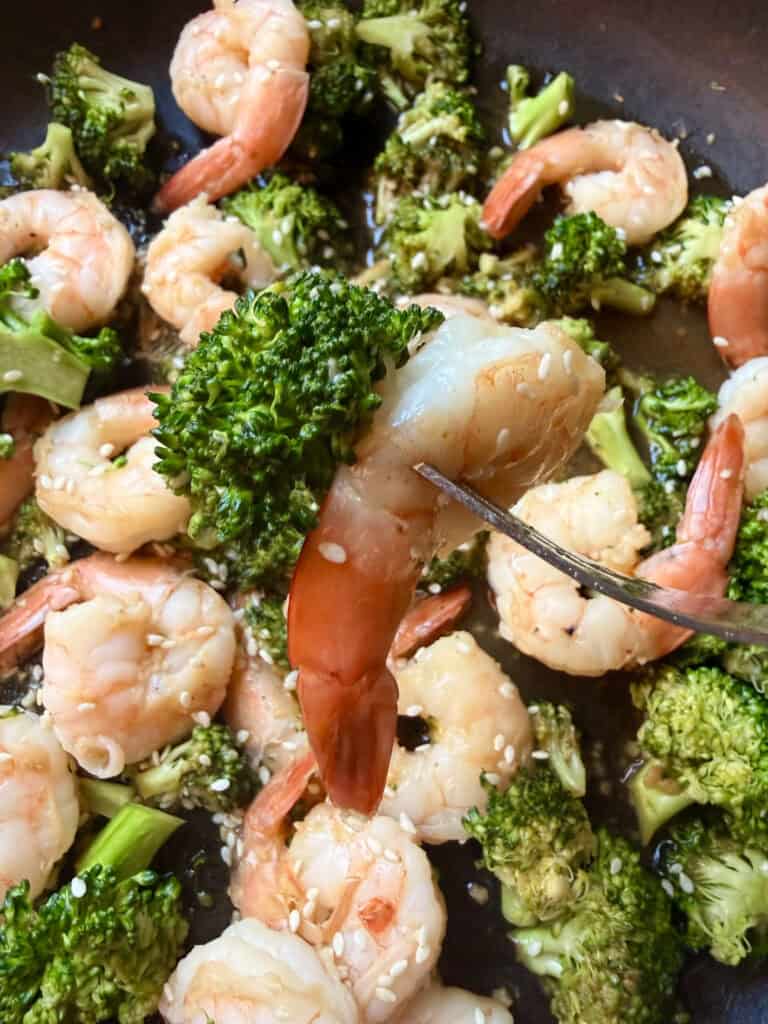 Close-up of a fork lifting a shrimp and broccoli from a skillet, coated in garlic sesame sauce and topped with sesame seeds.
