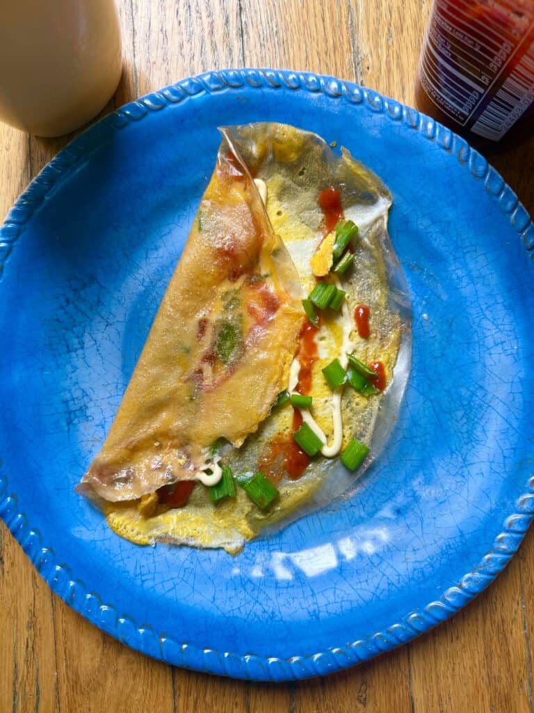 Low-carb egg wrap folded in half on a bright blue plate, topped with sriracha, mayonnaise drizzle, and chopped green onions.