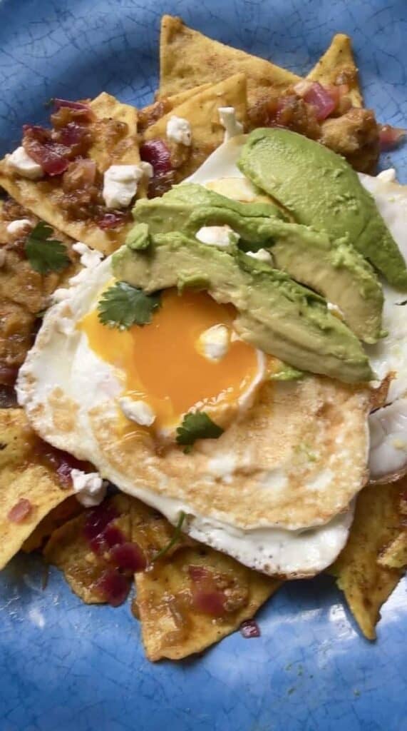 Chilaquiles verdes with salsa verde chips topped with a fried egg, avocado slices, queso fresco, cilantro, and diced red onion on a blue plate.