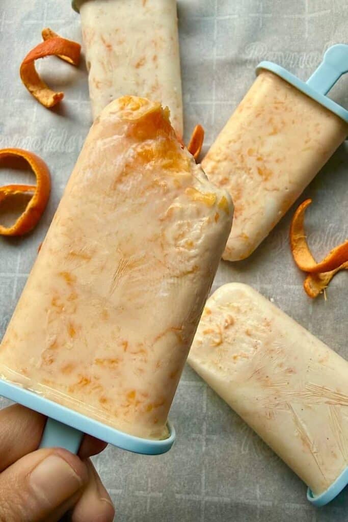 Hand holding a bitten orange creamsicle popsicle made with Greek yogurt and Cuties with frozen popsicles in the background