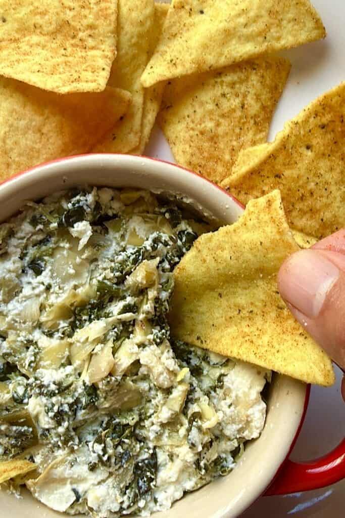 Quest tortilla chip dipping into baked spinach artichoke dip with Greek yogurt in a small ramekin”