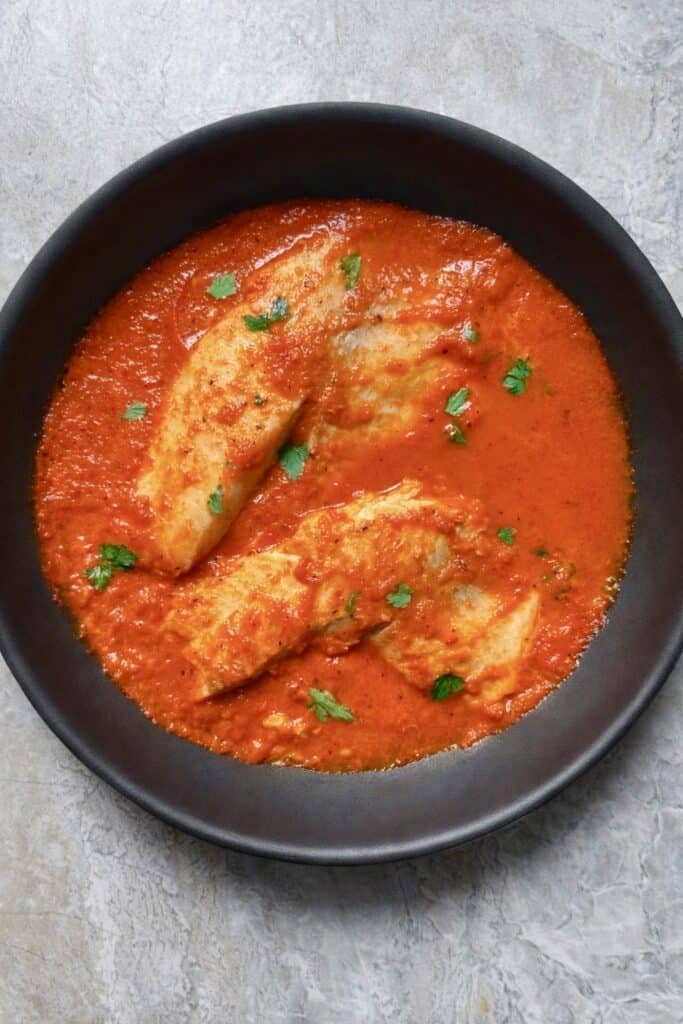 Top-down view of white fish fillets in a rich red pepper tomato sauce in a black skillet, garnished with parsley on a light stone background.