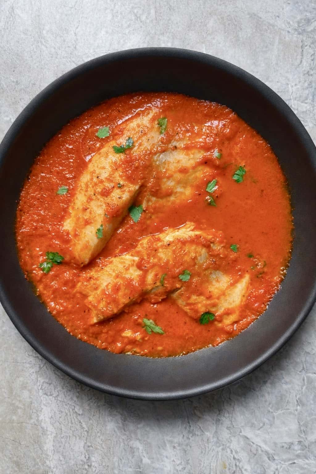 Top-down view of white fish fillets in a rich red pepper tomato sauce in a black skillet, garnished with parsley on a light stone background.