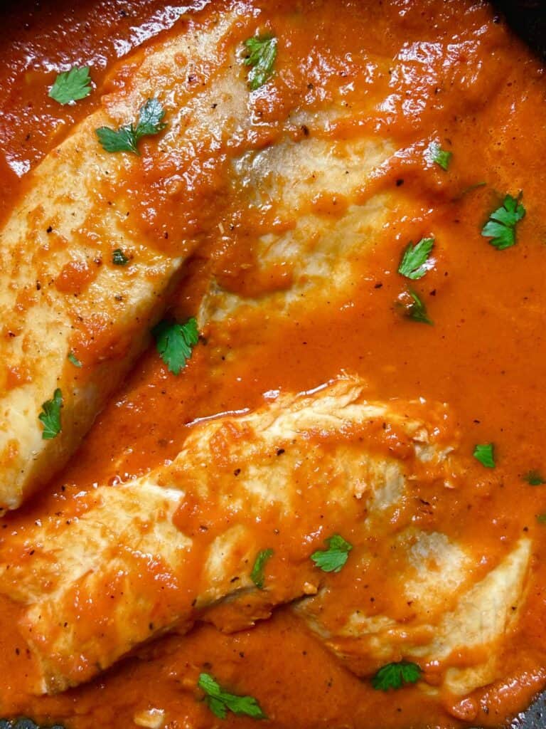 Close-up of pan-seared white fish fillets simmering in a creamy red pepper tomato sauce, garnished with fresh parsley.