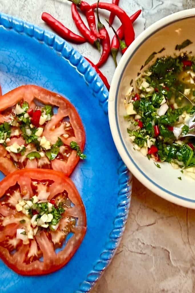 Crack sauce spooned over sliced tomatoes with chopped garlic, Thai chilies, and cilantro next to dipping bowl”