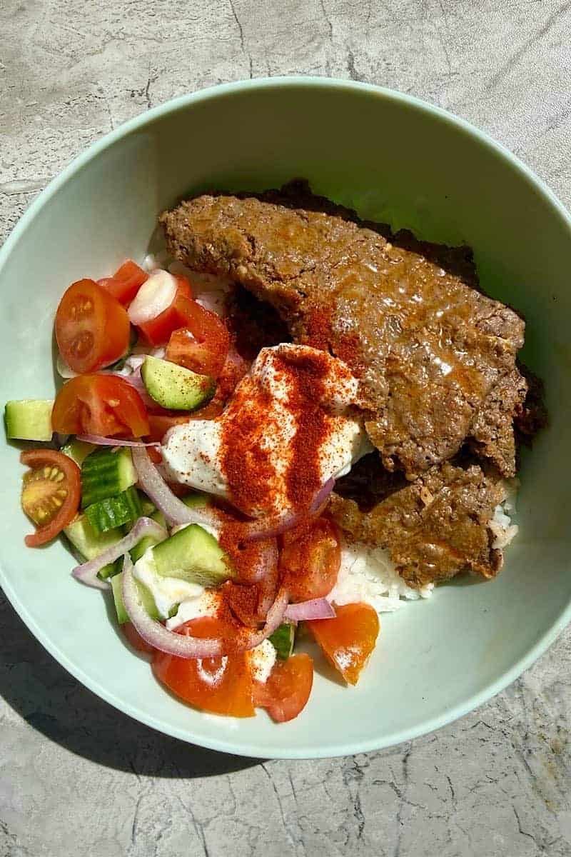 Homemade doner kebab bowl with ground beef slices, rice, yogurt sauce, paprika, tomato cucumber salad, and red onion