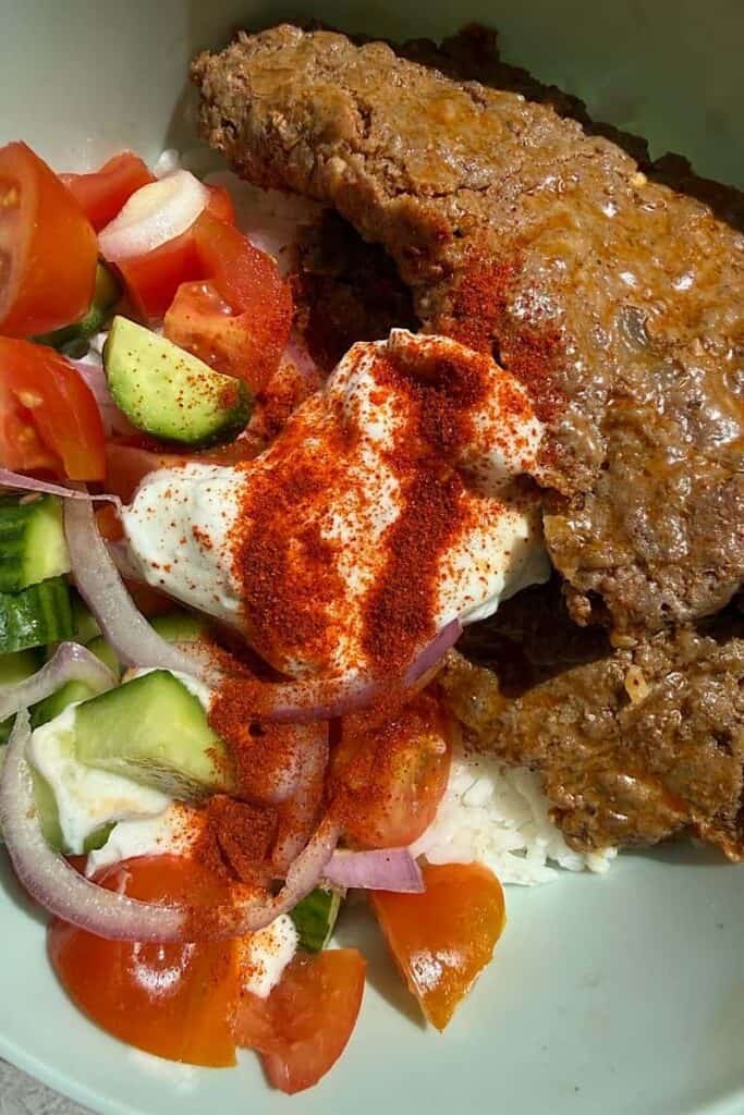 Close-up of homemade doner kebab bowl with ground beef, yogurt sauce, paprika, tomatoes, cucumber, and onions over rice
