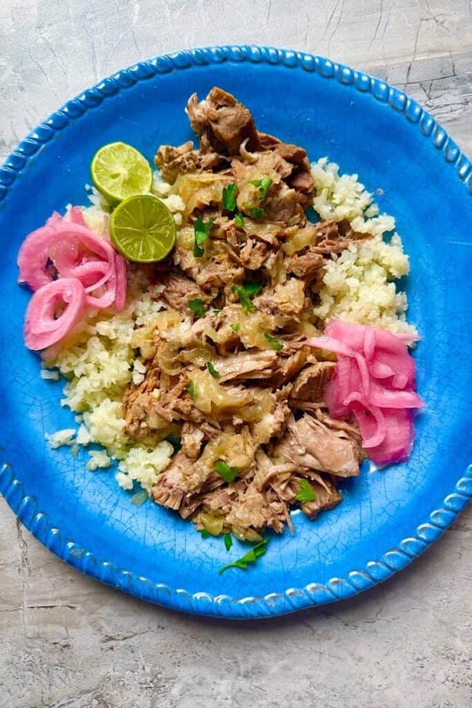 Instant Pot mojo pork served over cauliflower rice with lime wedges, pickled red onions, and fresh herbs on a blue plate