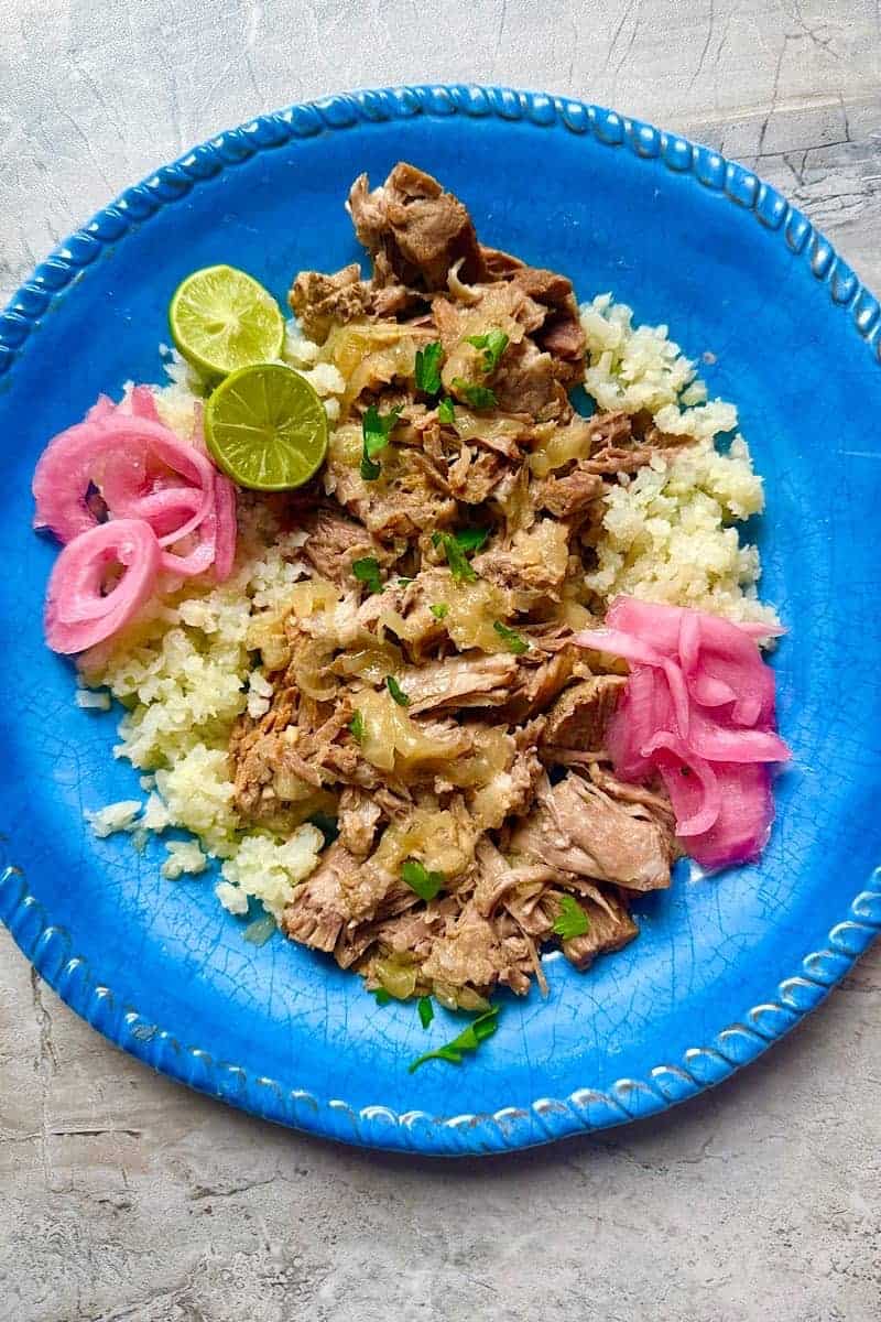 Instant Pot mojo pork served over cauliflower rice with lime wedges, pickled red onions, and fresh herbs on a blue plate
