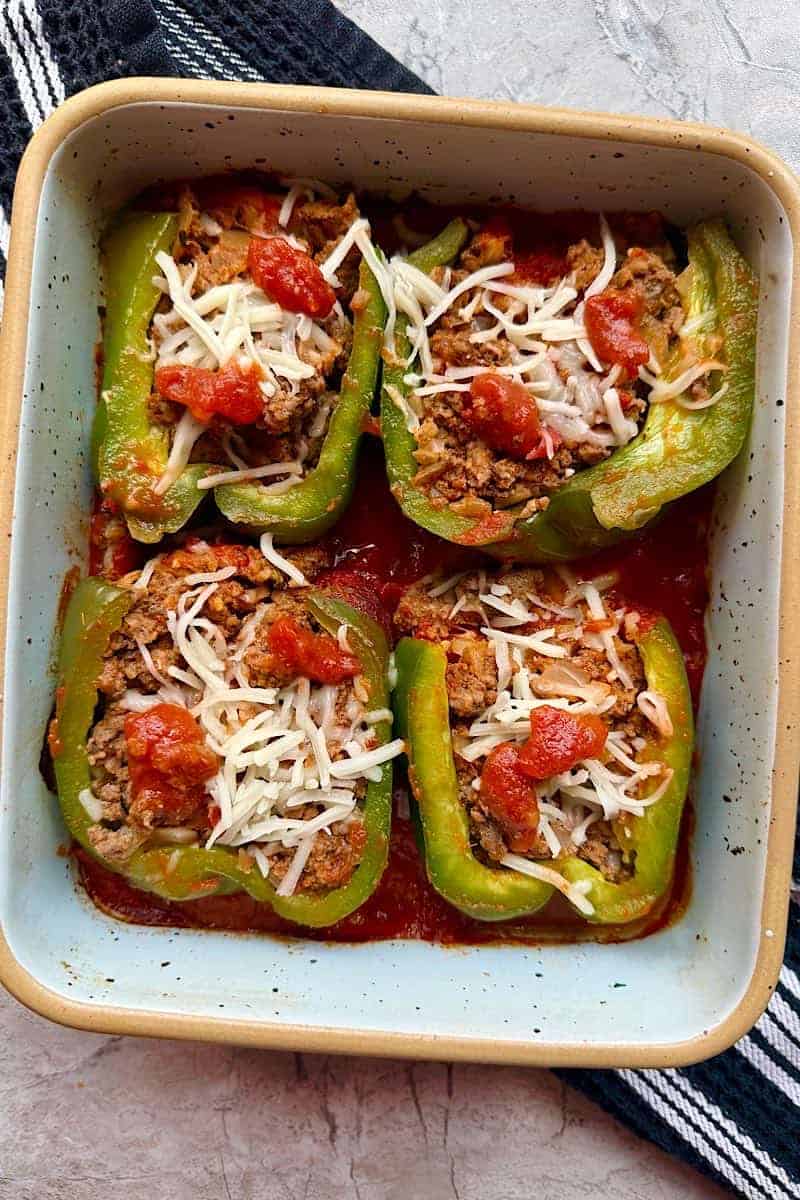 Close-up of baked stuffed green bell peppers filled with ground beef mixture, shredded white cheese, and dollops of red sauce, arranged in a light blue ceramic baking pan