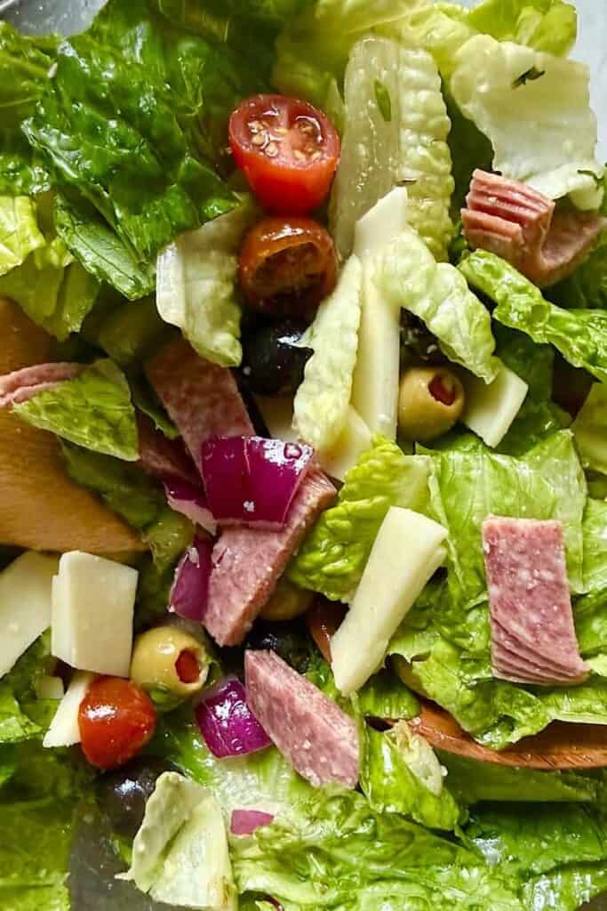 Close up of Italian chopped salad tossed in a glass bowl with romaine lettuce, salami, black and green olives, provolone cheese, and cherry tomatoes