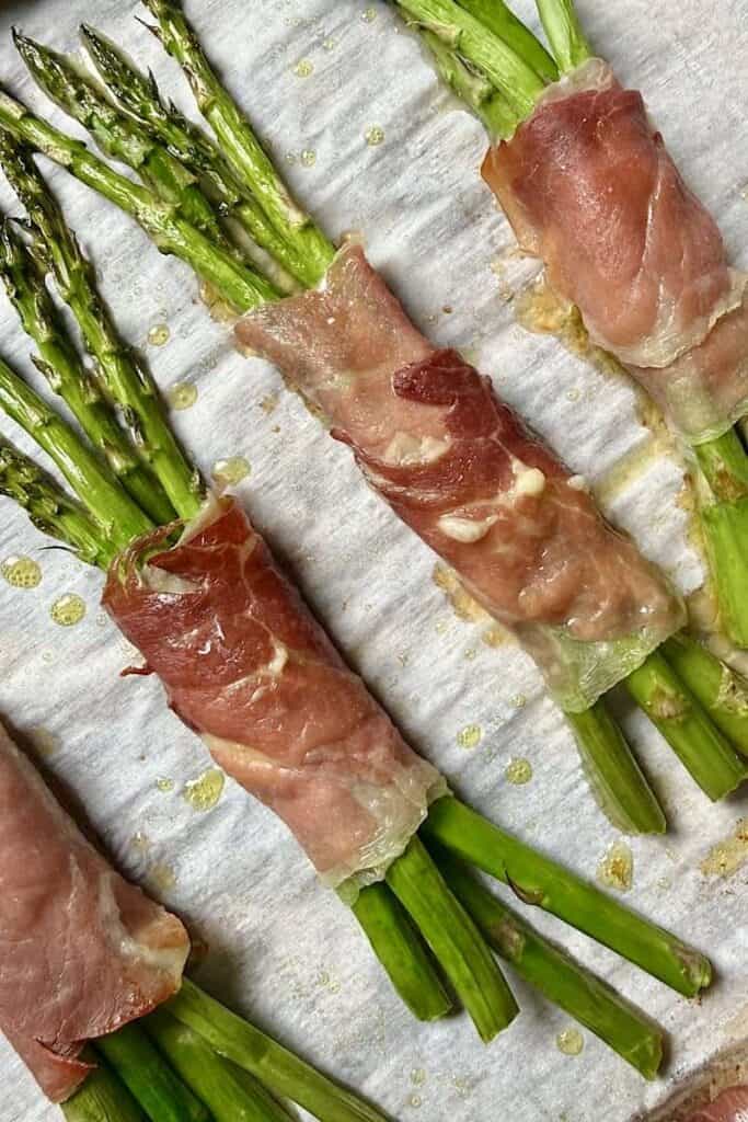 Prosciutto wrapped asparagus bundles baking on a parchment-lined sheet pan