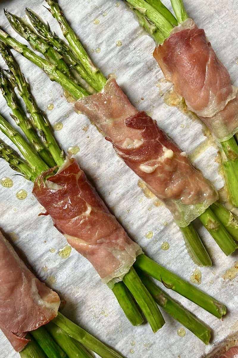Prosciutto wrapped asparagus bundles baking on a parchment-lined sheet pan