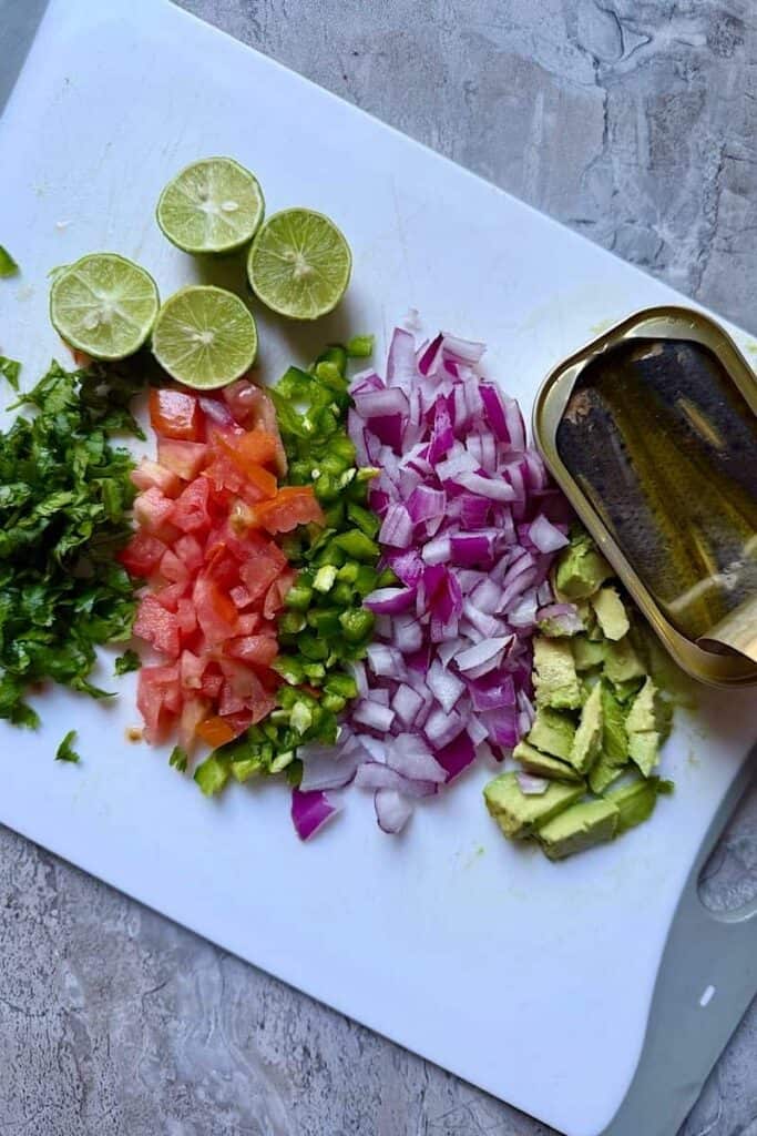 Chopped ingredients for sardine ceviche including tomatoes, red onion, jalapeño, cilantro, avocado, and fresh lime with canned sardines on a cutting board