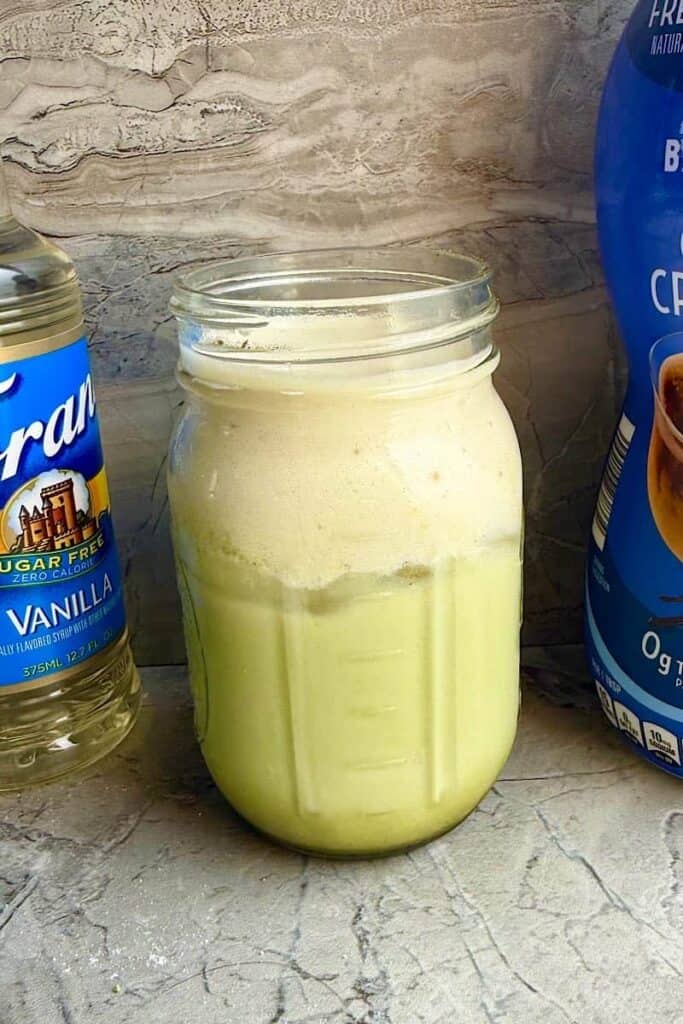 Iced banana bread matcha topped with banana cream cold foam, served beside sugar-free vanilla syrup and French vanilla coffee creamer.